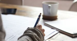 person writing on brown wooden table near white ceramic mug